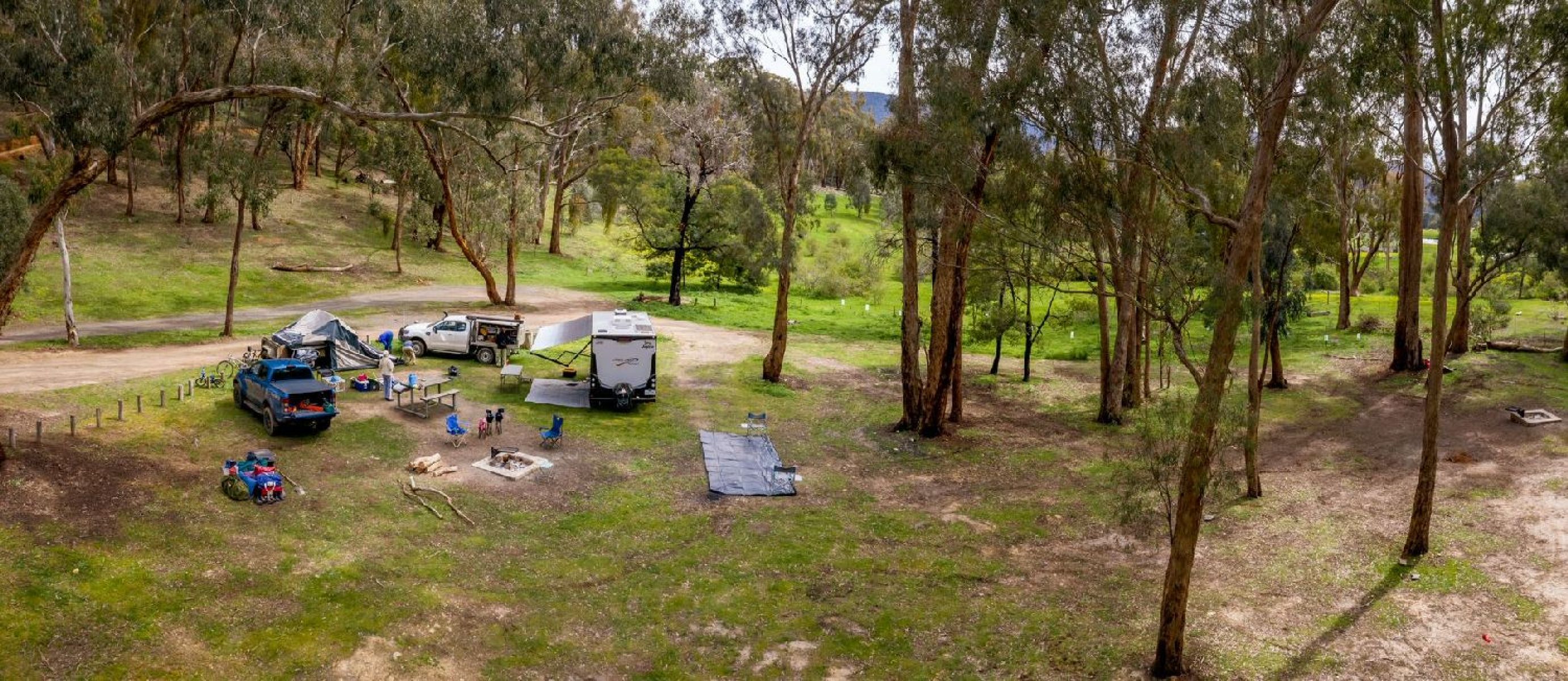 Cars and a campervan parked at a camping site in the forest, setting up tents and camp chairs around the fire