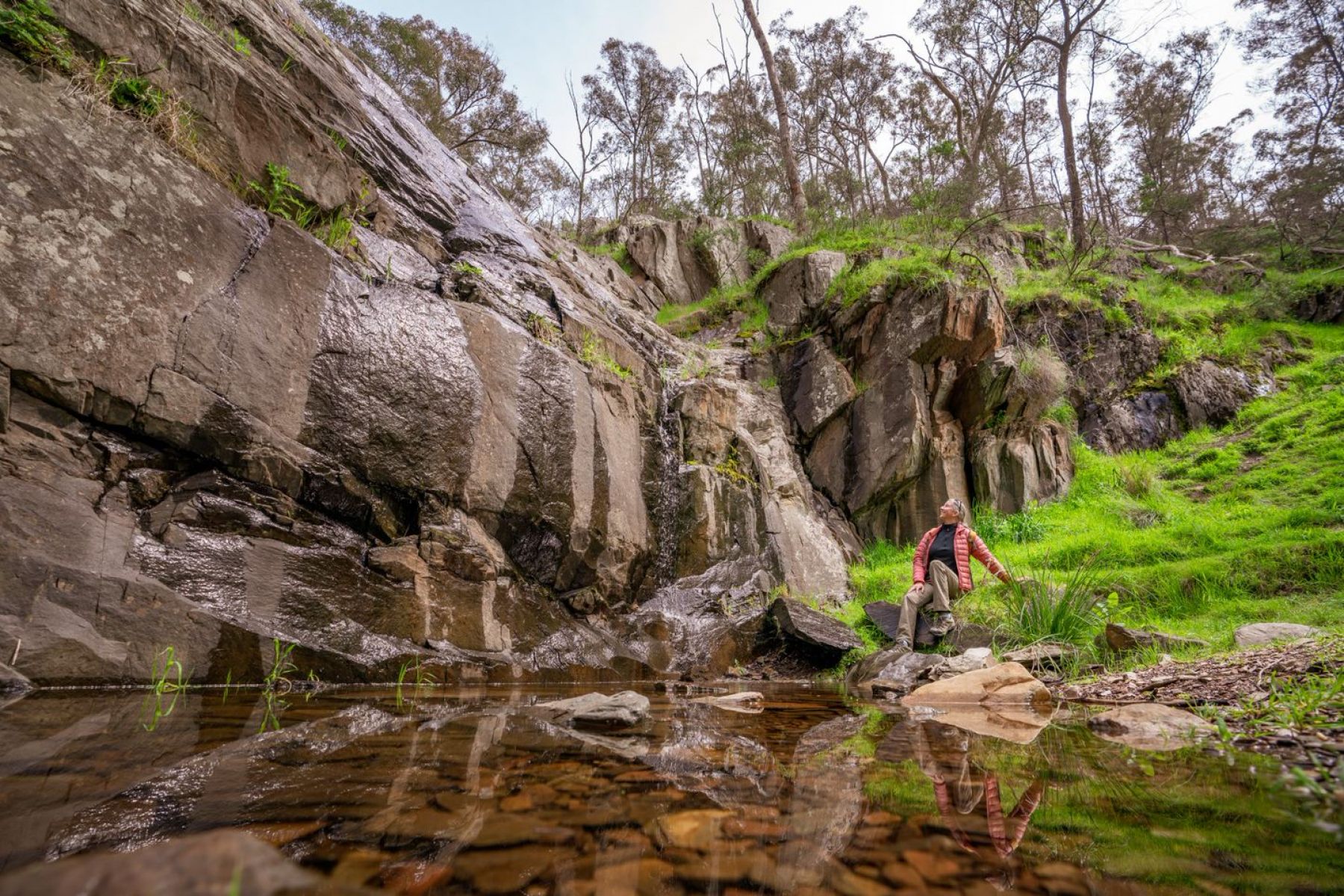 A person sitting and looking up at a large rock face, with water from Avoca falls pouring down