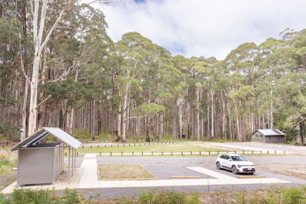 A wide, flat footpath runs between a gravel carpark, an open grassy area and a toilet block.