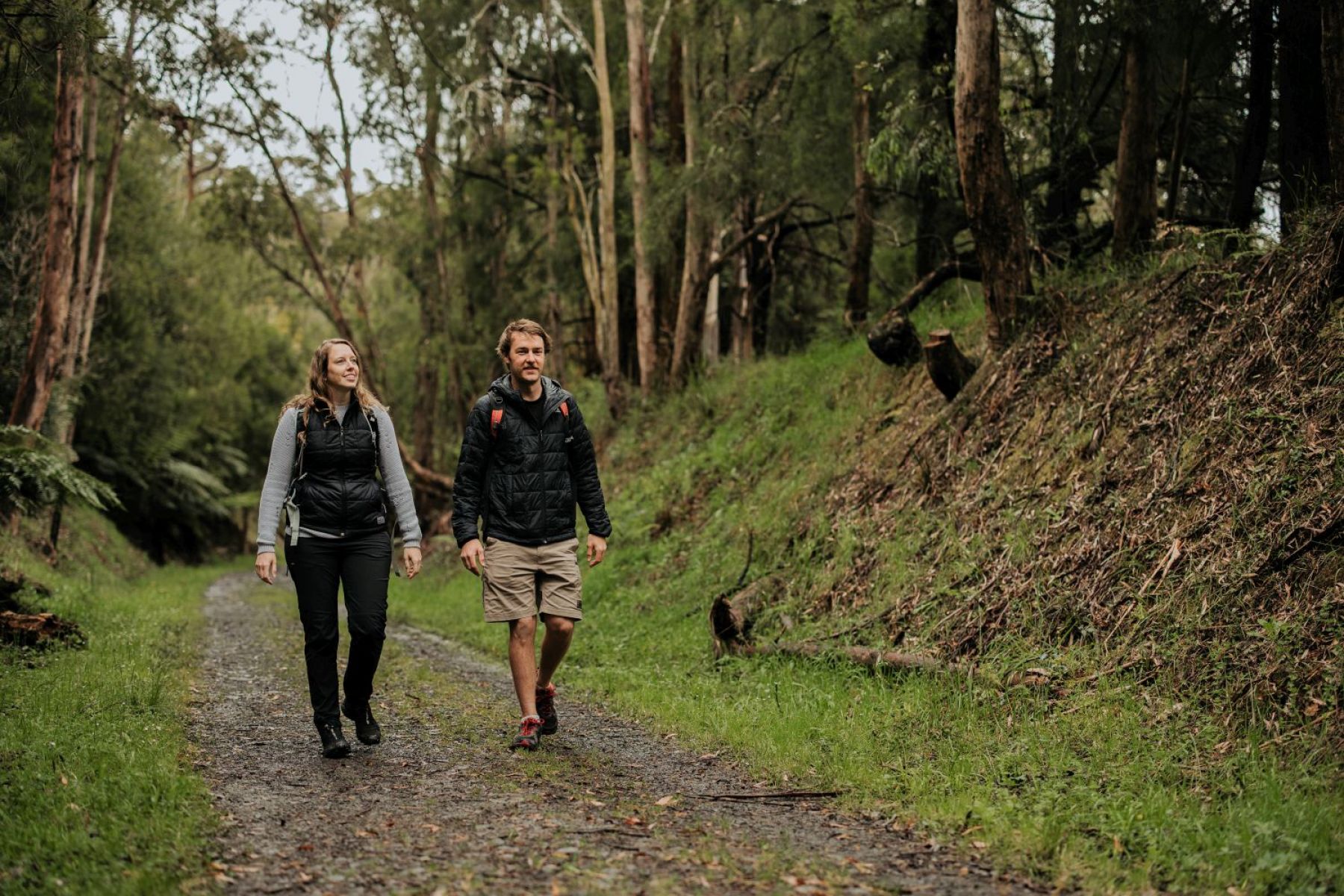 Two people walking on the trail. They are walking on a dirt trak with tall trees behind them.