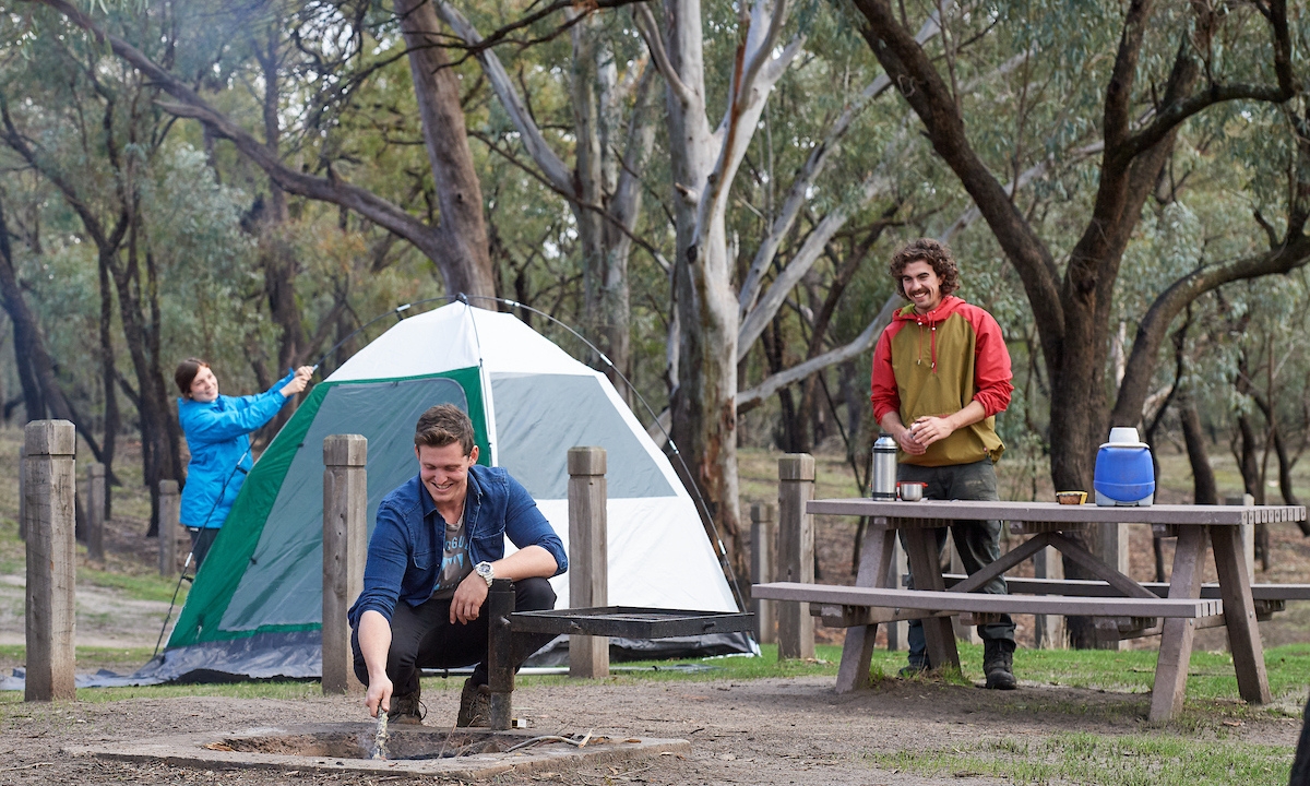 A ground of friends set up their campground. A tent is in the background, a man crouches down by a firepit and another mn stands behind a picnic table pouring himself a drink. 