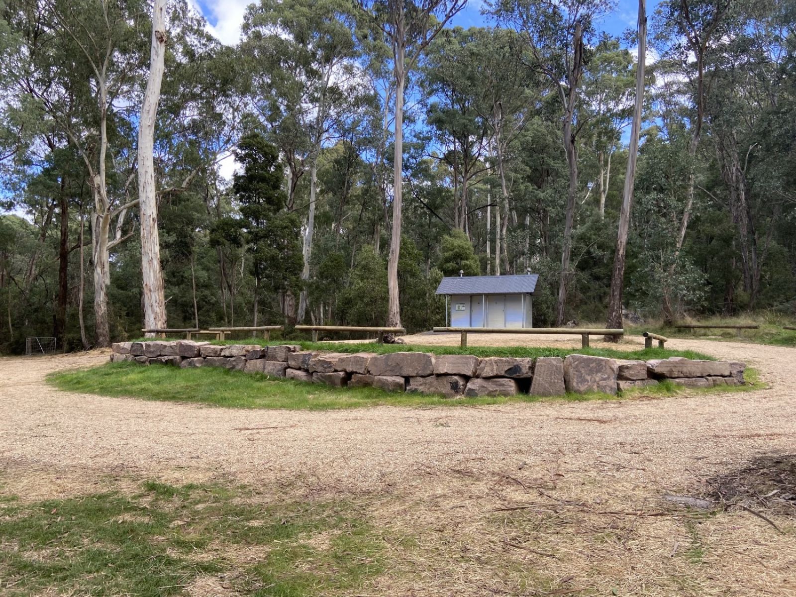 A dirt road circles around a small patch of grass and some boulders. Behind this road is a metal toilet block.