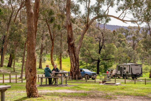 People sitting on a picnic bench eating lunch, with their cars in the background and people setting up a tent