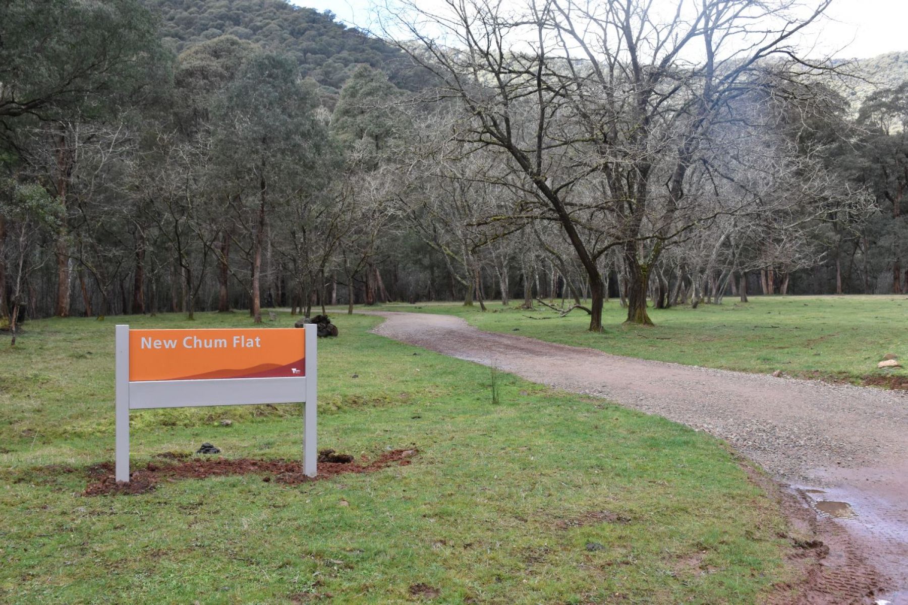 An orange entry sign next to a road which leads into a flat, green and grassy campground with hills in the background. campground.