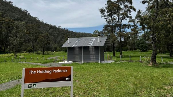 A sign reading The Holding Paddock' sits in front of a green grassy camping area and a metal toilet block. Tree-covered mountains are in the background. 