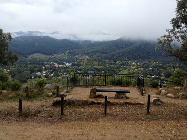 A wooden bench at a lookout with views of a town below and cloud-covered mountains in the distance