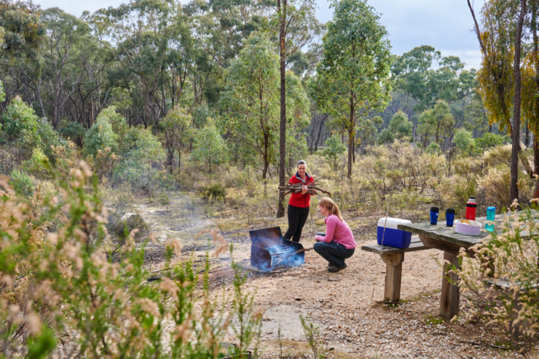 Two women load wood into a firepit in the bush. 