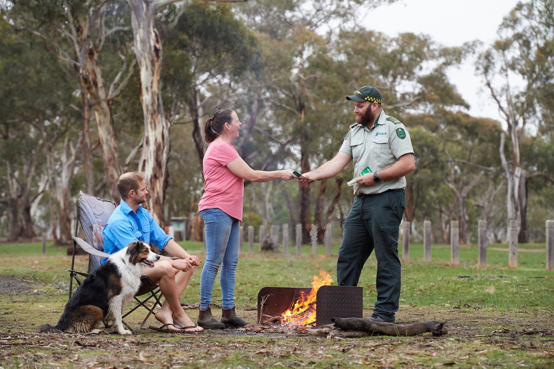 A man in an FFMVic uniform speaks to two people sitting with their campfire. They are all smiling. 