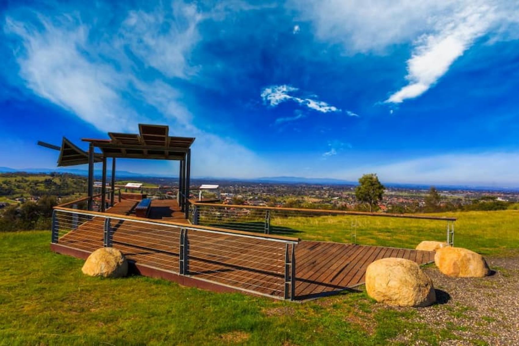 A wooden lookout platform and shelter with metal railings, overlooking the north-eastern suburbs of Melbourne with blue skies and light clouds. There are boulders at the walkway onto the platform