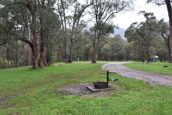 Gravel road through the campground, with campsites on either side and an amenity block at the end. Green grass and a fire pit in the foreground. 