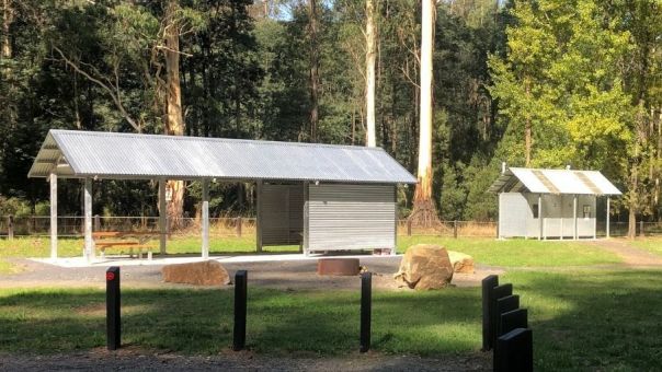 A metal picnic shelter and table next to a toilet block in an open grassy areas surrounded by trees.