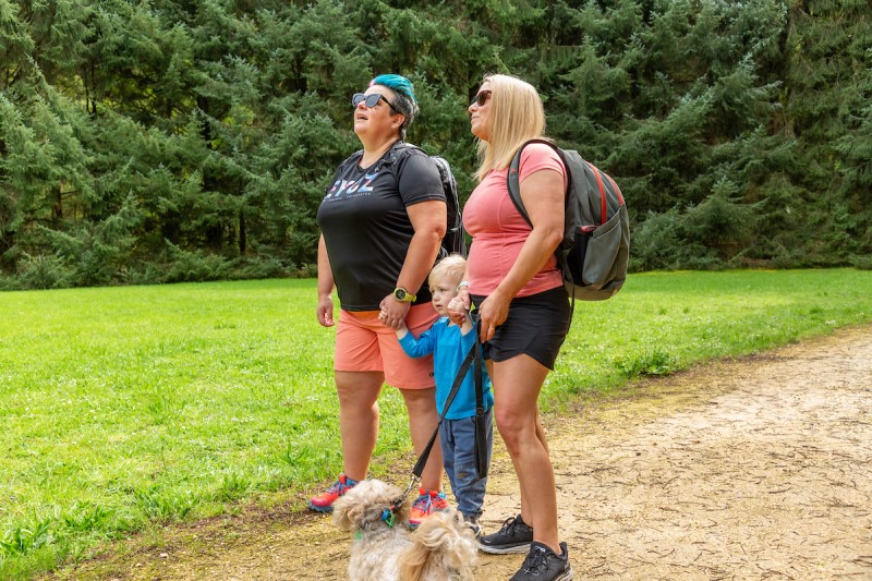 Two women and a child with their dog look ahead with tall green pines behind them on a walking trail.  