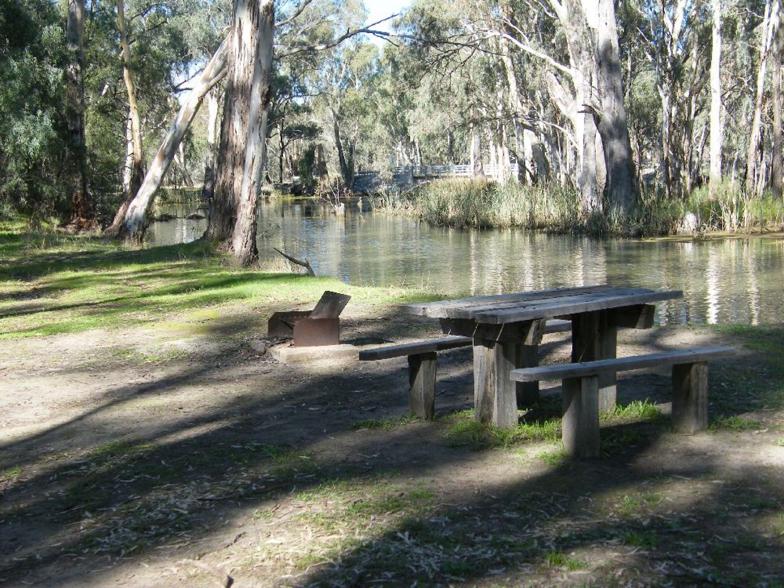 A picnic bench on the grassy banks of the river with the bridge in the background