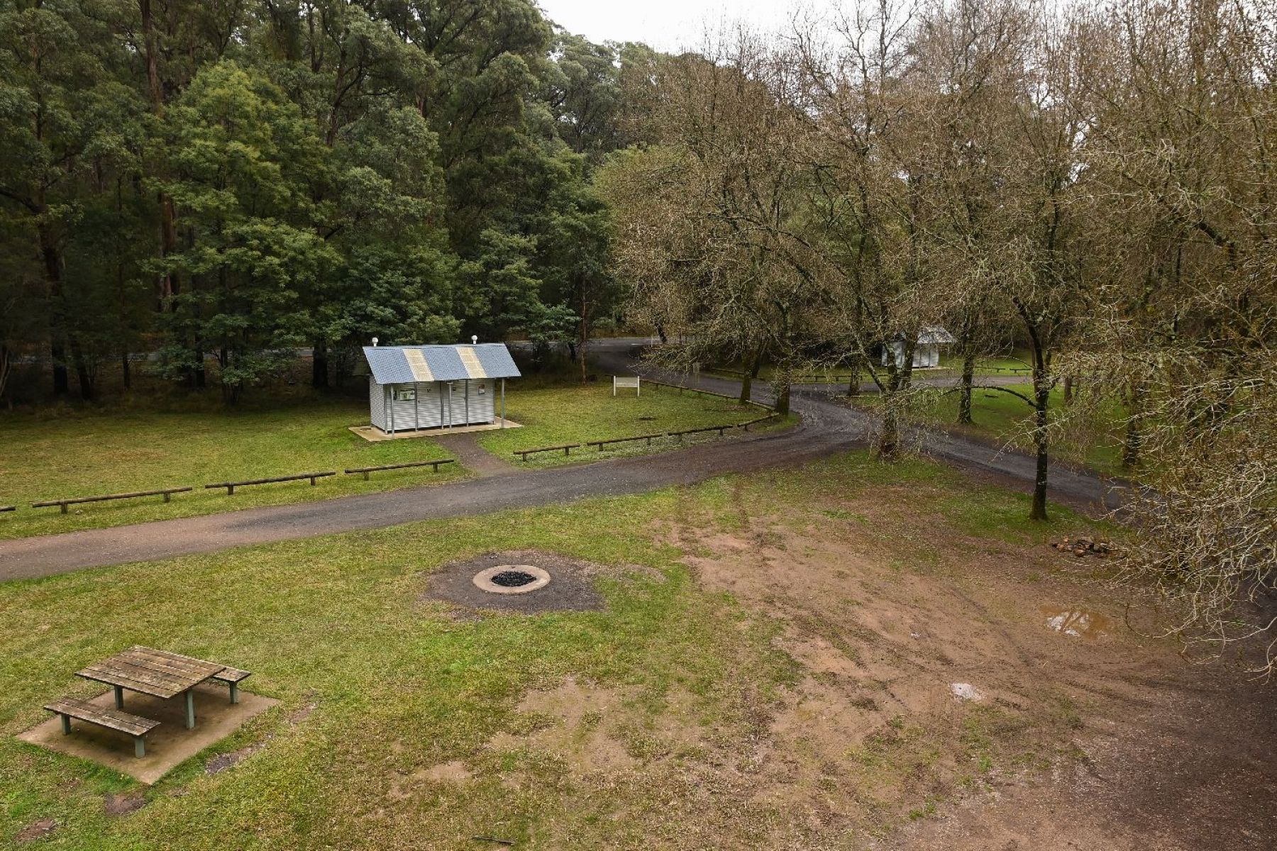 Vew of the wide open camping area with plenty of space. A firepit, table and chairs with a building in the background. Mature trees behind the small building.