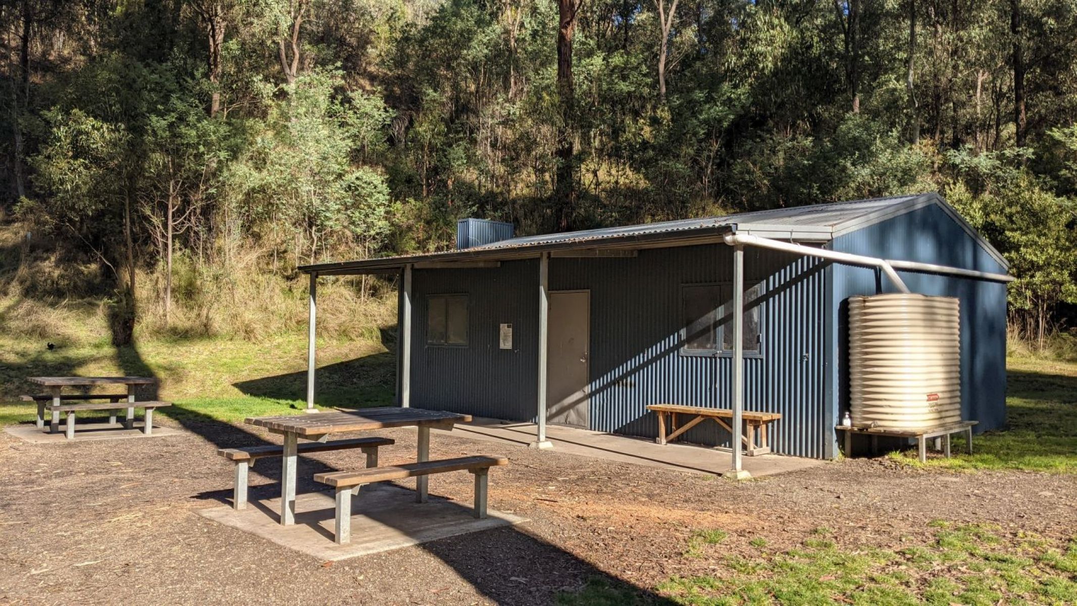 Two wooden picnic tables, a bush hut and an adjoining water tank.