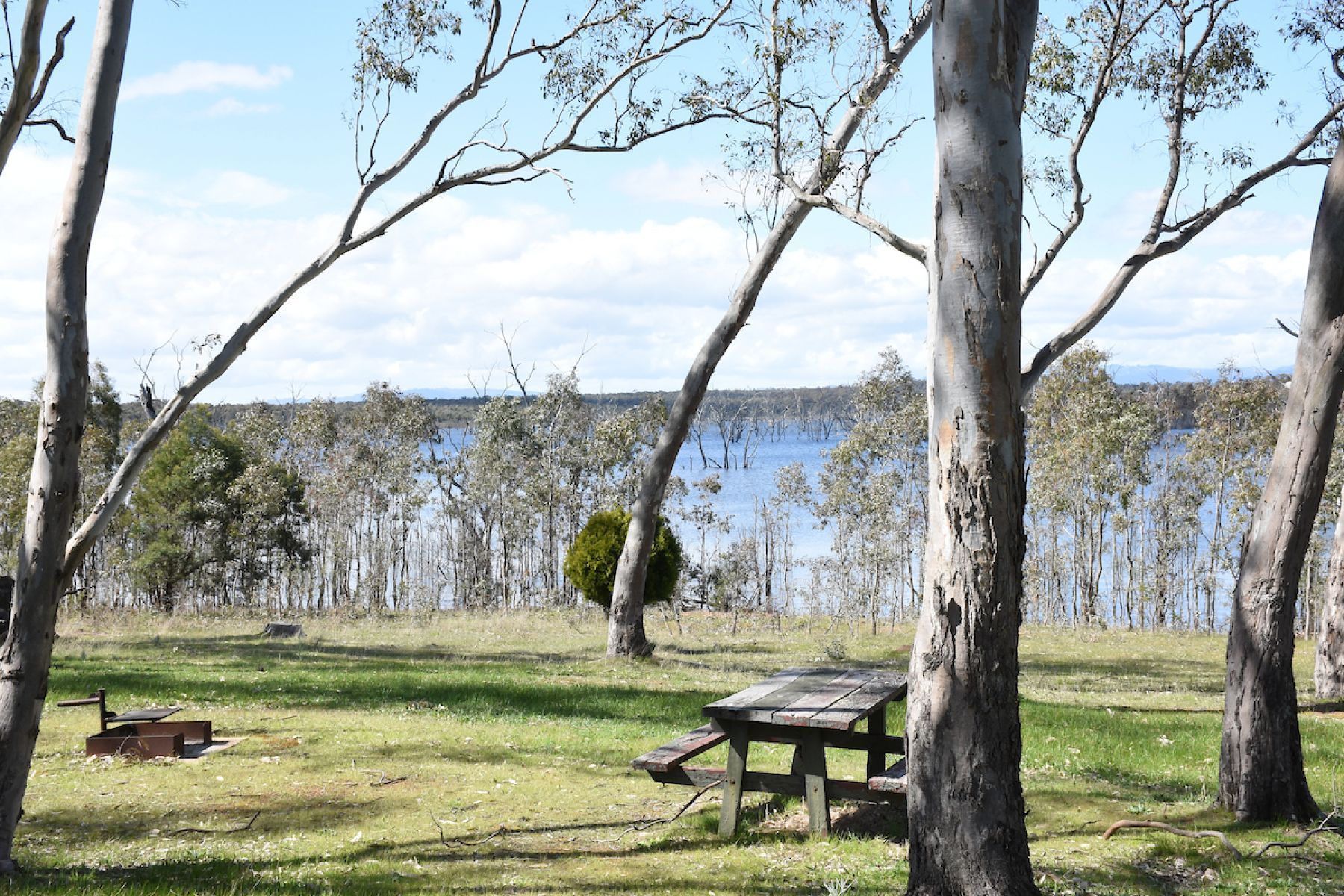 Grassy camping area with a wooden camping table, fire pit and gum trees overlooking a lake. 