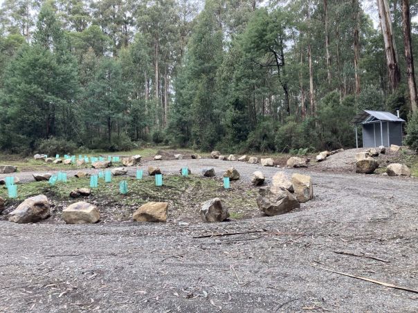 A flat gravelled area bordered by boulders and a metal toilet facility to the side. A variety of trees surround the area. 