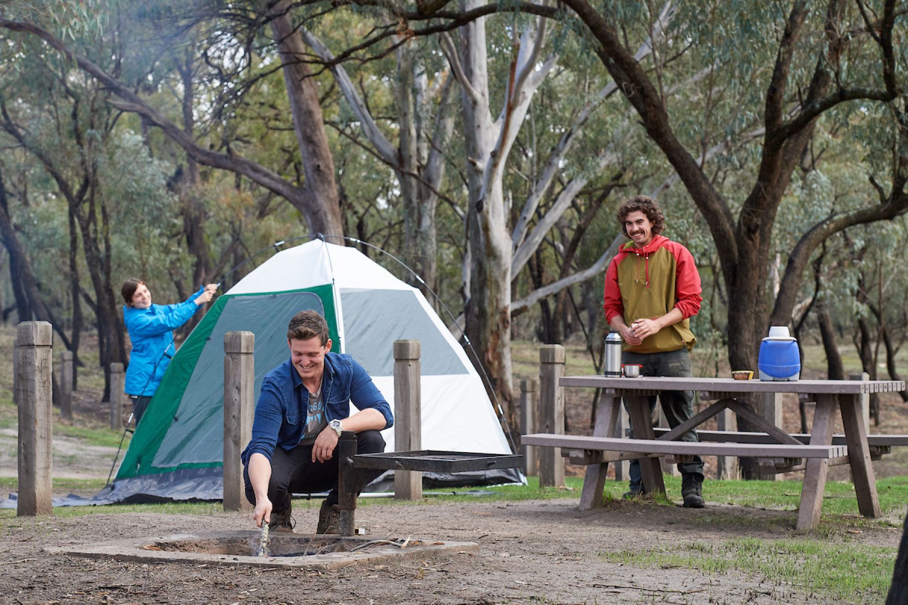 A ground of friends set up their campground. A tent is in the background, a man crouches down by a firepit and another mn stands behind a picnic table pouring himself a drink. 