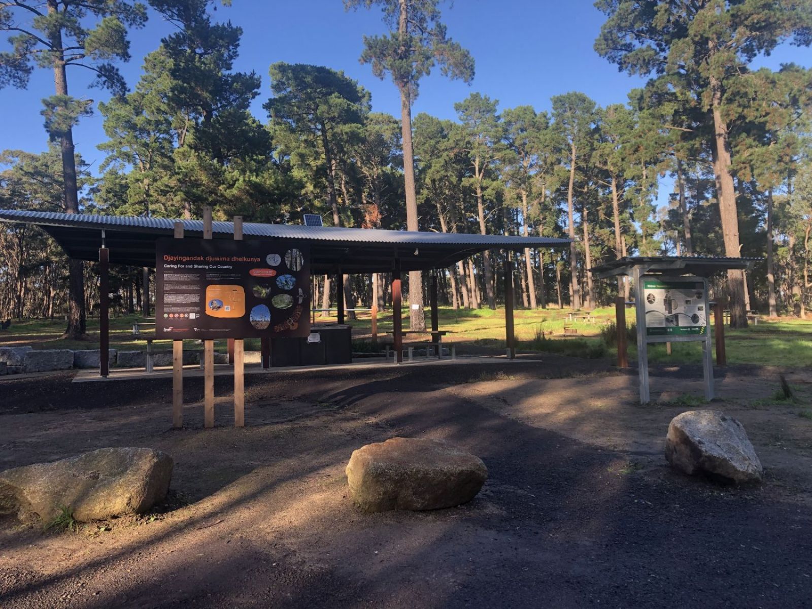 Large rock bollards border a path in foreground, with information signage boards and a picnic shelter in the background. The site is surrounded by tall pine trees.