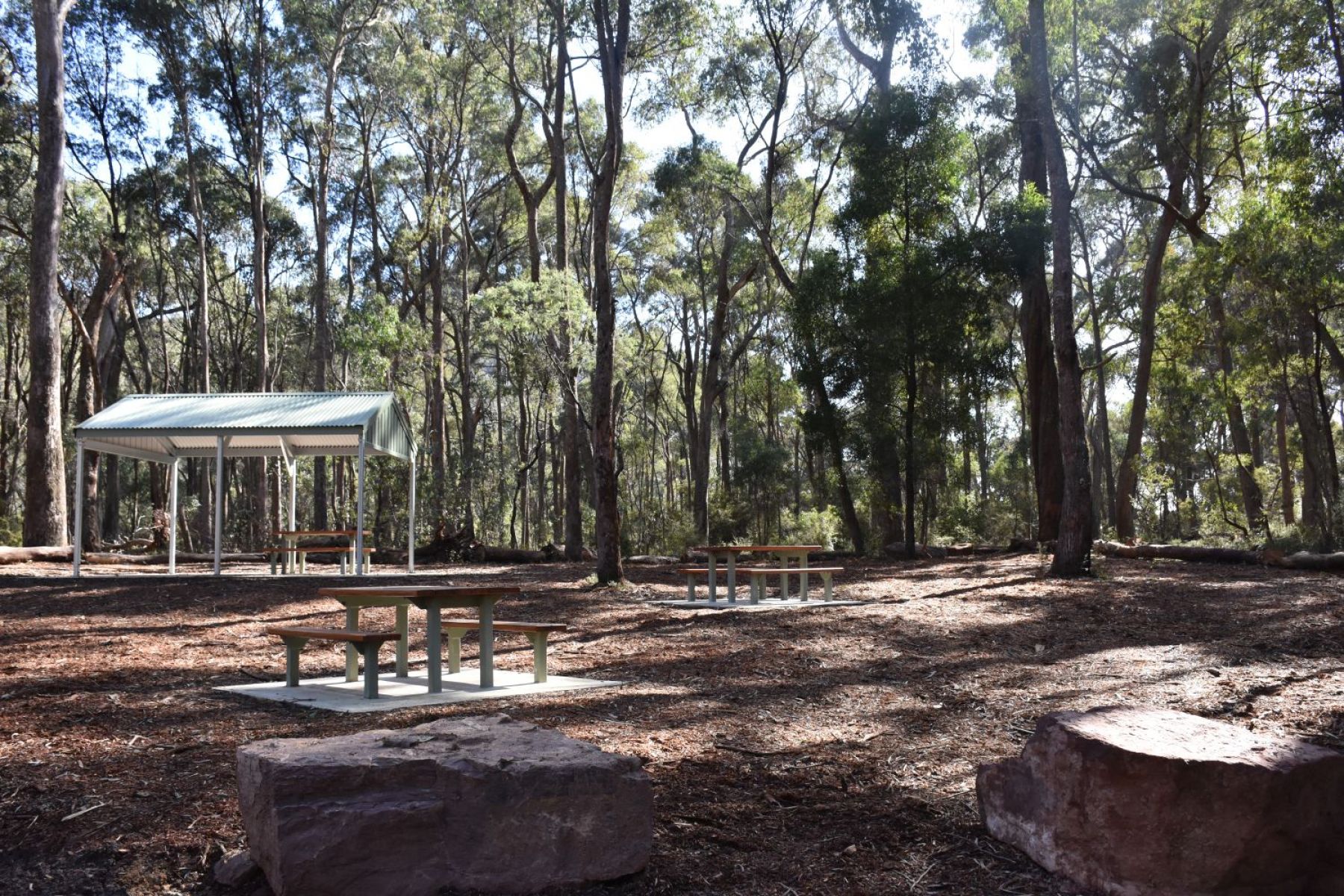 A wooden picnic table sits in front of a picnic shelter shaded by tall leafy trees,