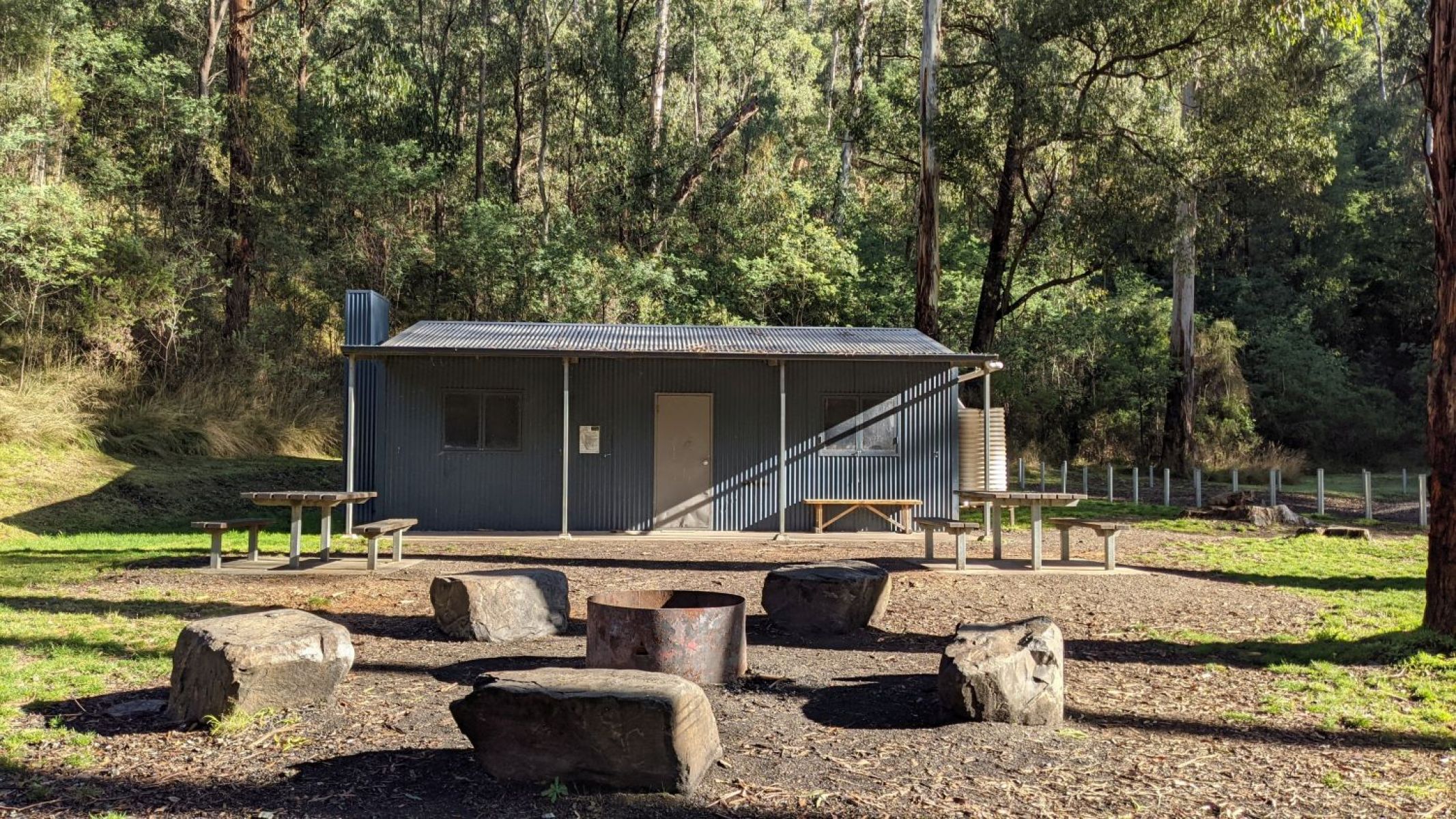 Seats made from rock surround a metal firepit. A metal hut is in the background and shady trees are behind it. 