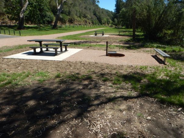 A wooden picnic table next to a metal fire pit. A gravel road and another picnic table are in the background surrounded by bushland.