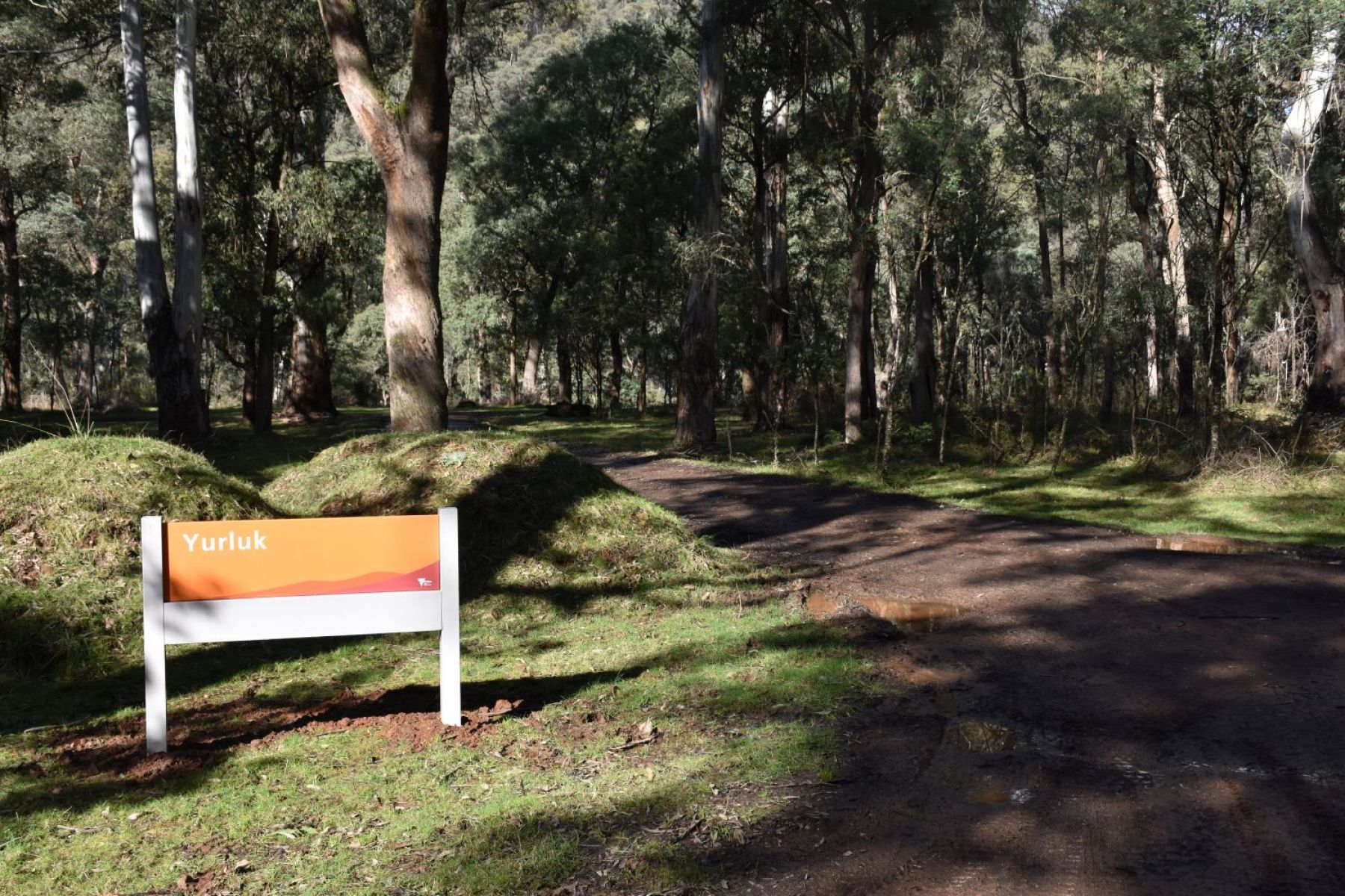 An orange sign reading Yurluk next to a dirt road leading into a bush campground. 